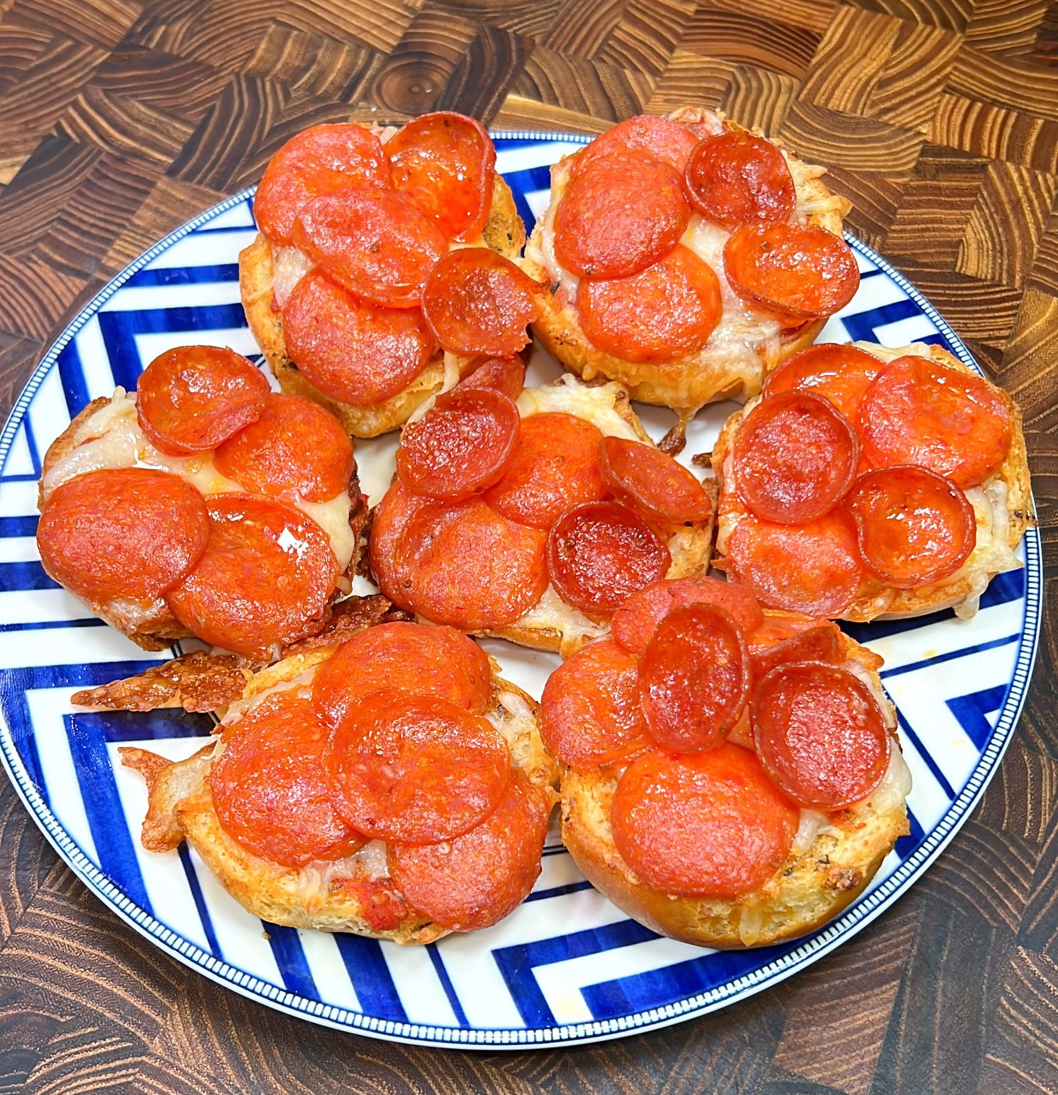 A plate holds six mini pizzas, each topped with melted cheese and slices of pepperoni. The pizzas are arranged on a wooden table and served on a blue and white patterned plate.