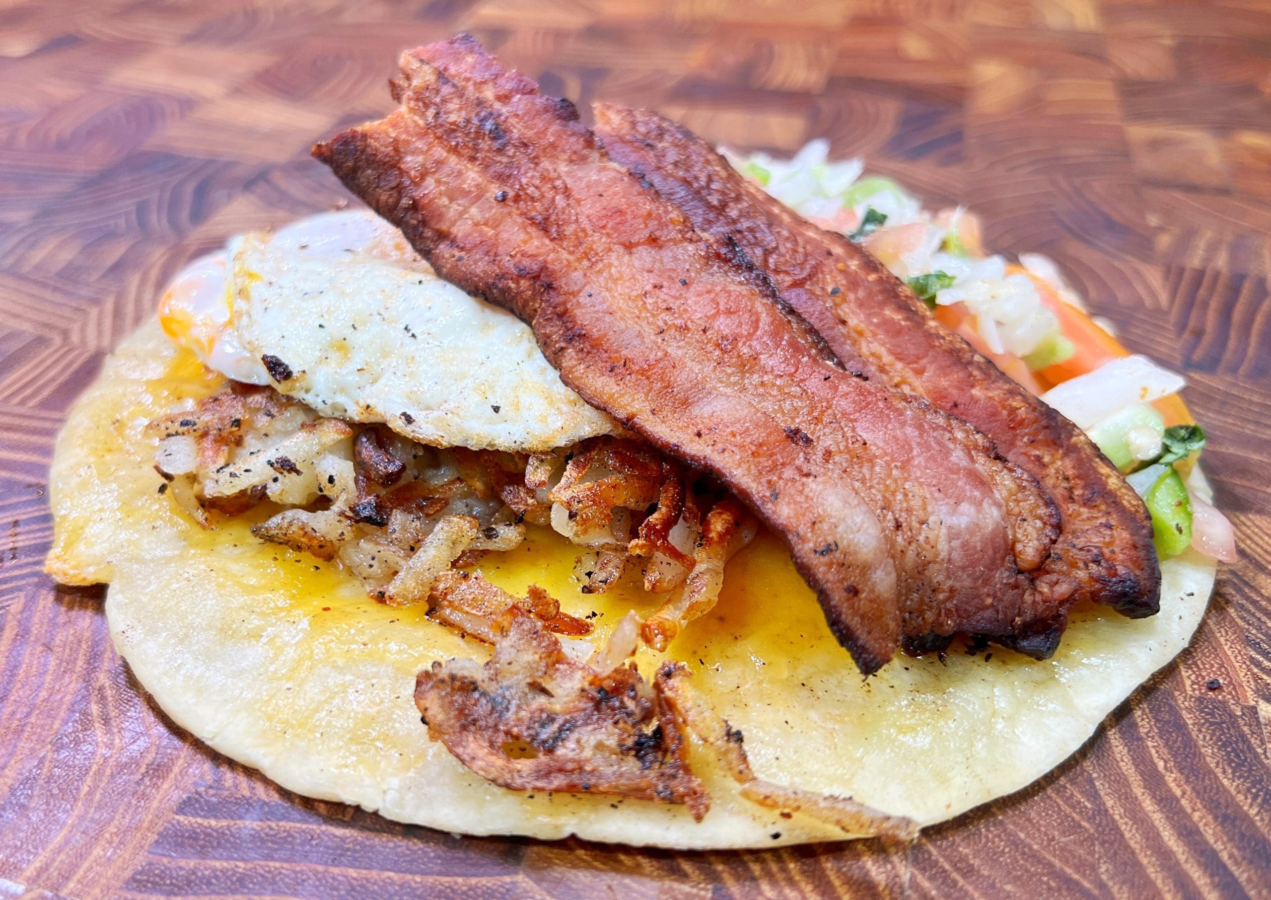A breakfast taco with a crispy corn tortilla topped with grilled bacon strips, a sunny-side-up egg, and seasoned hash browns, served with a side of diced tomatoes, onions, and green peppers on a wooden surface.