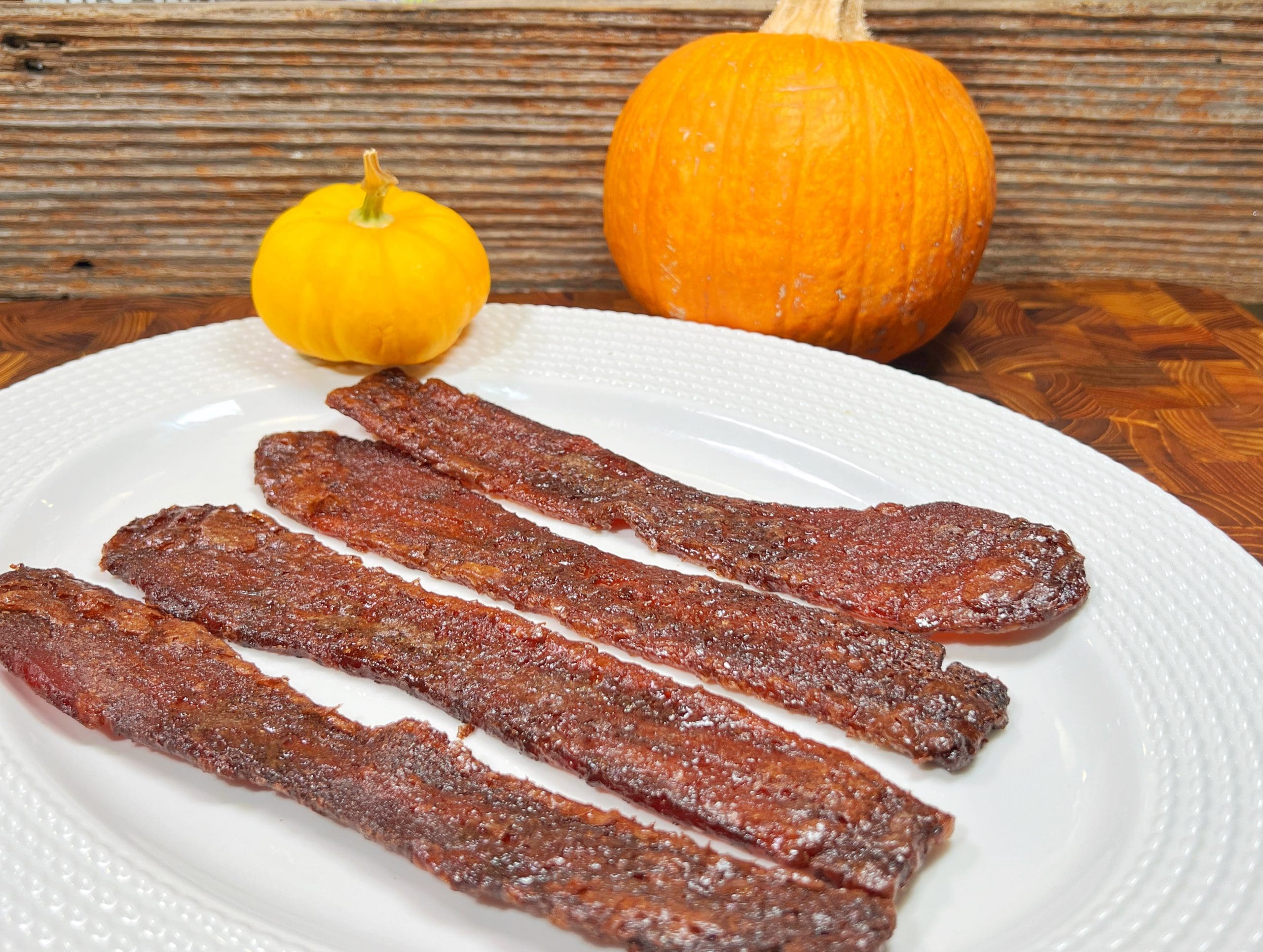 A plate with four strips of crispy bacon, alongside a small yellow gourd and a medium-sized orange pumpkin, set against a wooden background.