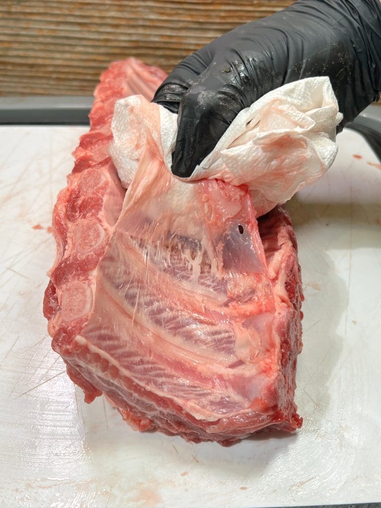 A hand in a black glove uses a paper towel to remove the membrane from a rack of raw ribs on a white cutting board.