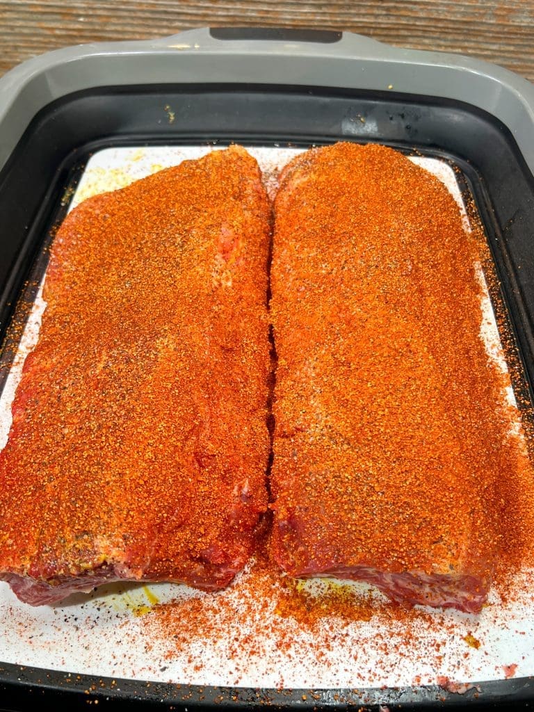 Two slabs of seasoned meat are placed on a tray, generously coated with a reddish-brown spice rub. The background features a textured wooden surface.