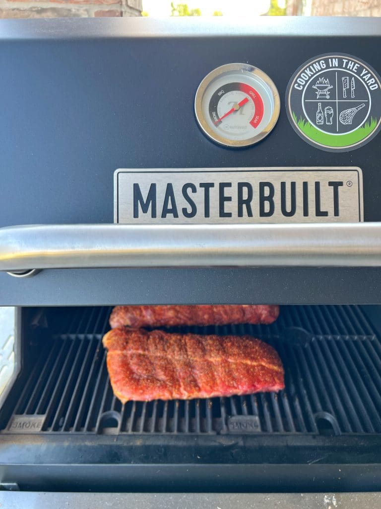 Masterbuilt grill with seasoned ribs cooking inside. The grill lid features a temperature gauge and a sticker that reads Cooking in the Yard.