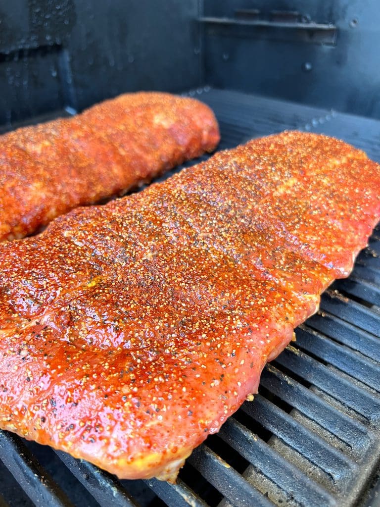 Two racks of pork ribs coated with a reddish seasoning blend are grilling on a barbecue. The seasoning appears to include a mix of spices, creating a textured look. The ribs are sizzling over clean, blackened grill grates.