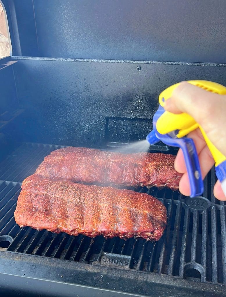 A person is spraying a mist onto two racks of ribs on a grill. The ribs are seasoned and cooking, with the grill lid open. The persons hand is holding a yellow and blue spray bottle, adding moisture to the ribs.