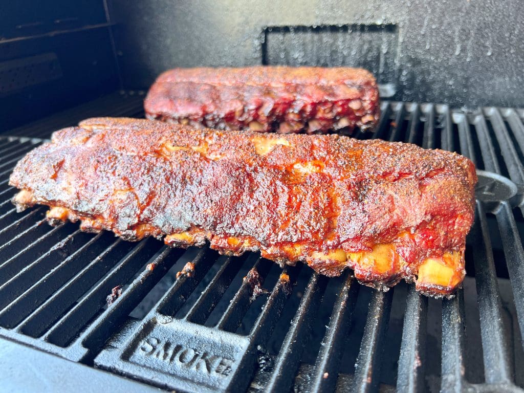 Close-up of two racks of barbecued ribs on a grill, with a rich, smoked glaze. The ribs are sitting on a black grill grate, displaying a crispy, caramelized crust. The background shows a textured, dark wall of the grill.