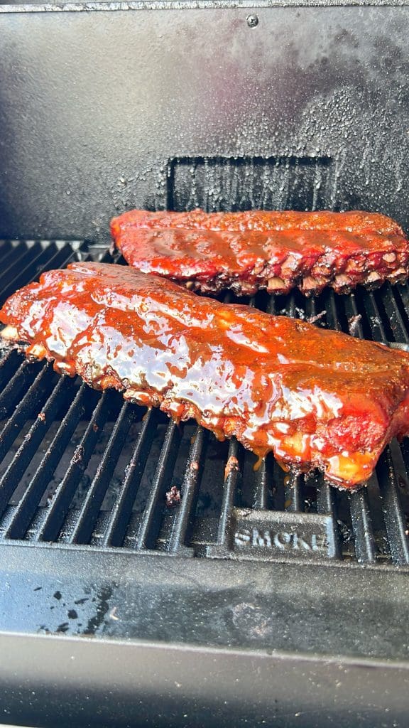 Two racks of smoked ribs coated in a glossy barbecue sauce cooking on a grill, with a smoky backdrop. The ribs are positioned horizontally and their rich, caramelized glaze is highlighted under natural light.