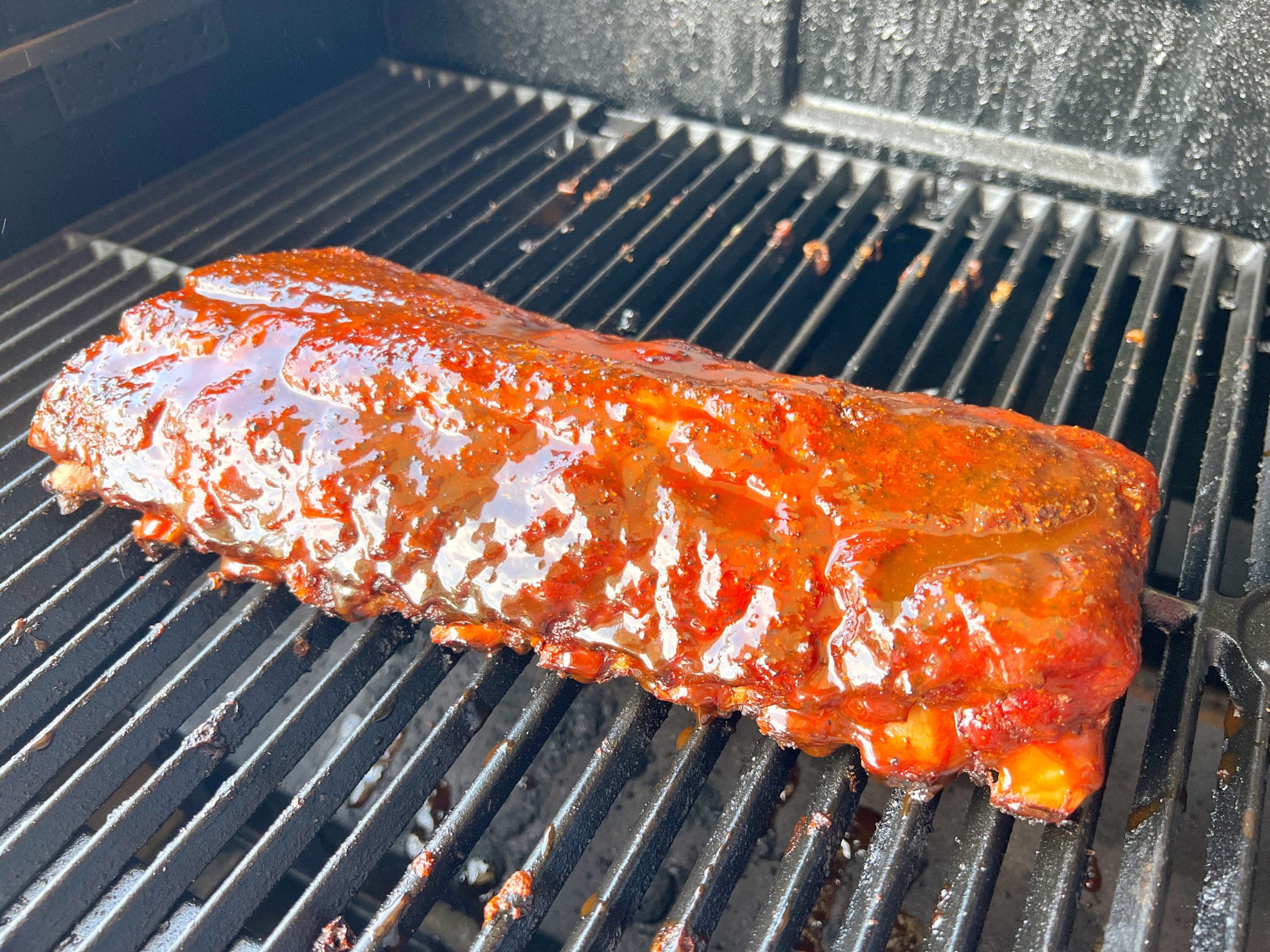 A rack of glazed barbecue ribs cooking on a black grill. The ribs are covered in a shiny sauce, highlighting their rich flavor and texture. The grills ridged surface adds to the mouthwatering appeal of the dish.