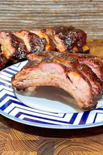 A plate of barbecued ribs with a glossy, caramelized finish on a wooden table. In the background, a larger portion of ribs is visible. The ribs are served on a plate with a blue and white zigzag pattern.
