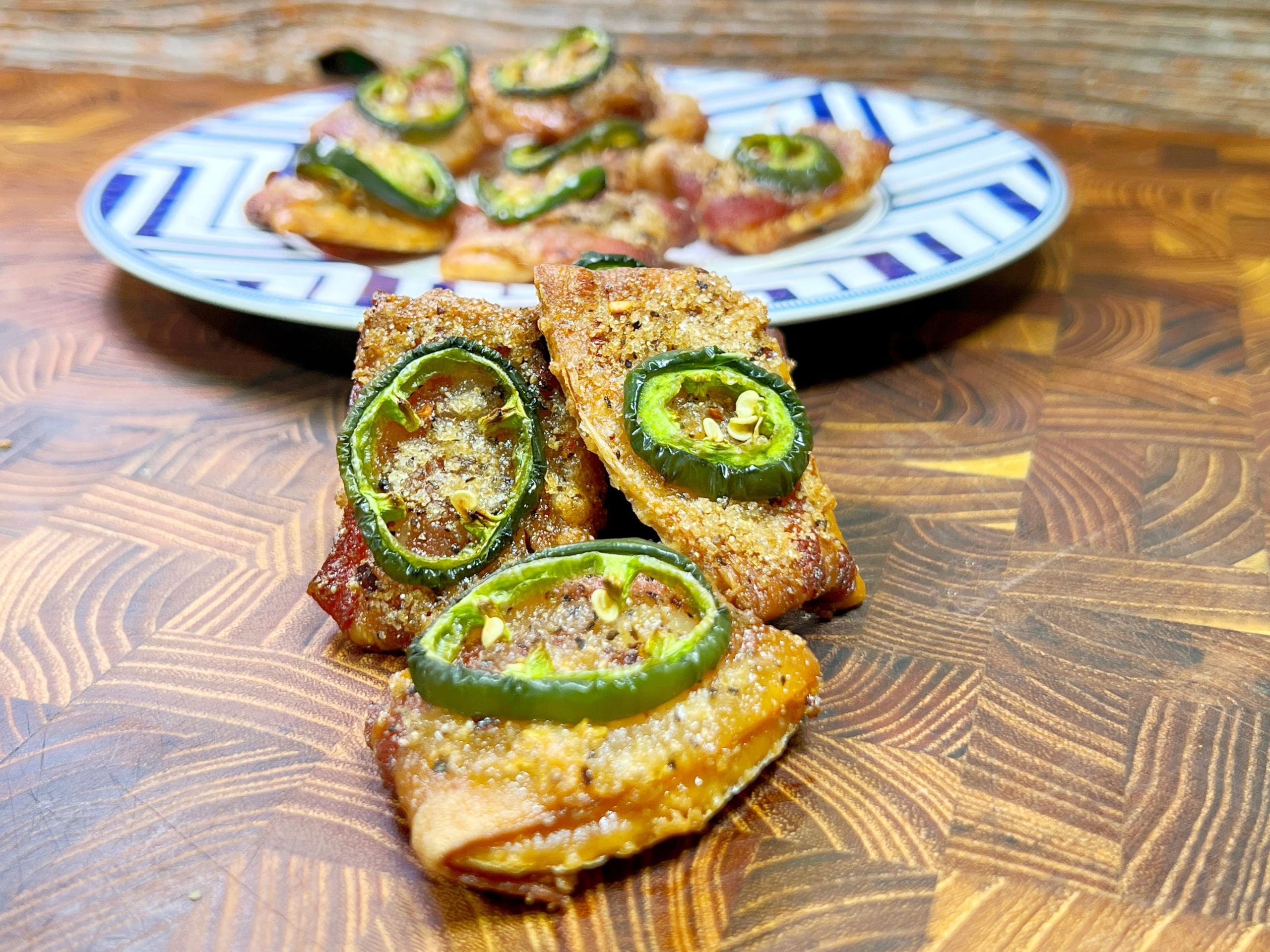 Close-up of spicy candied bacon wrapped crackers topped with jalapeño slices on a wooden table. A patterned blue and white plate with more candied bacon crackersis in the background.