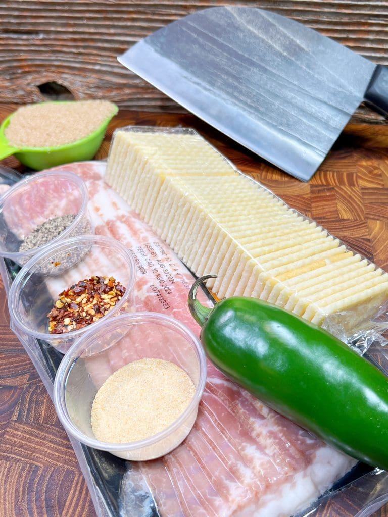 A wooden surface showcases ingredients for a spicy cheese and bacon dish: sliced cheese, a whole jalapeño pepper, a block of bacon, and small containers of red pepper flakes, black pepper, and garlic powder. Nearby lies a cleaver ready to prep Spicy Candied Bacon delights.