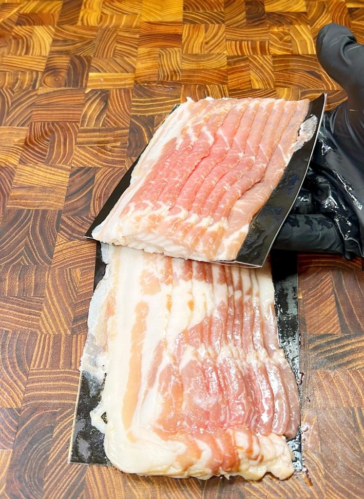 Sliced candied bacon is displayed on black packaging, resting on a wooden cutting board with a herringbone pattern. A gloved hand is partially visible to the right, adding a touch of spice to this delectable scene.