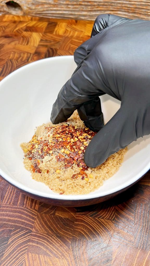 A gloved hand is mixing a spicy blend of brown sugar and red chili flakes in a white bowl, set on a wooden surface.