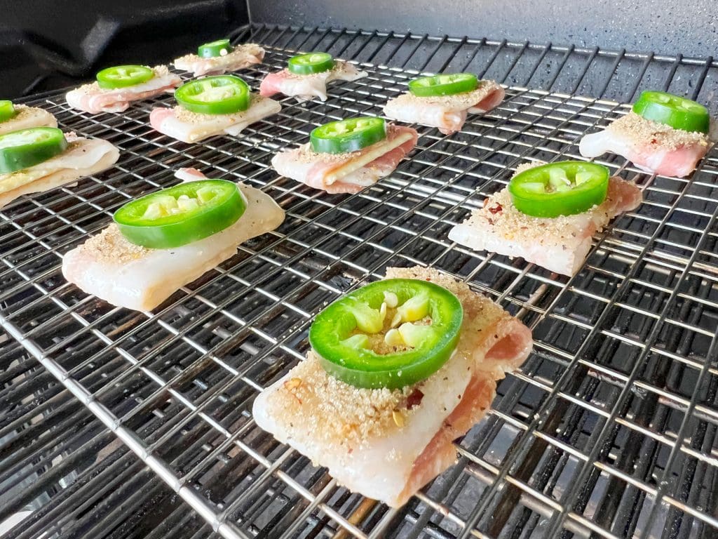 Slices of bacon-wrapped fish topped with jalapeño slices are arranged on a grill rack, ready to be cooked.