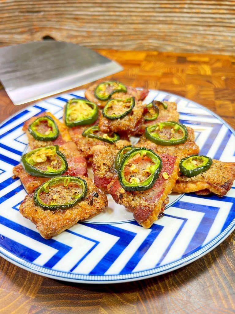 A plate of candied bacon wrapped jalapeño poppers rests on a zigzag-patterned dish. The poppers are topped with slices of fresh jalapeños, and a spatula is visible in the background on a wooden surface.