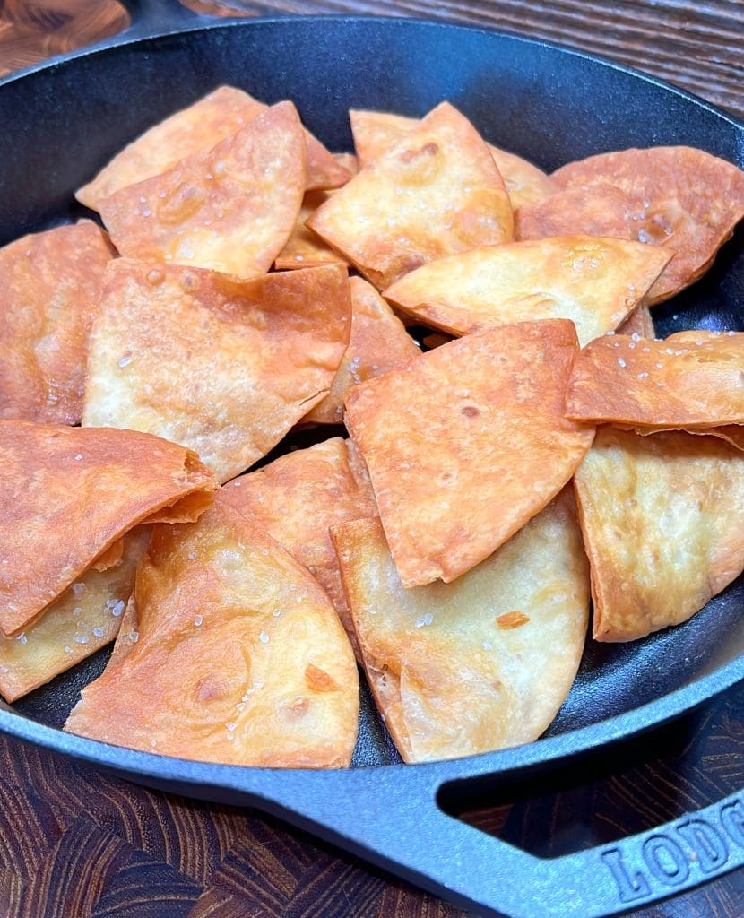 A close-up image of a cast iron skillet filled with golden-brown homemade tortilla chips, lightly sprinkled with salt. The chips are crisp and triangular, resting on a wooden surface.