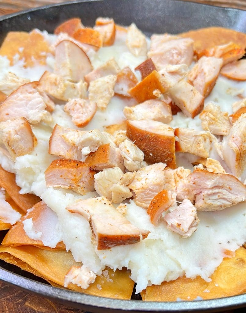 A close-up of a dish featuring grated cheese and chopped pieces of meat atop fried tortilla chips, arranged in a skillet. The cheese is melting slightly, and the tortilla chips provide a crispy base.