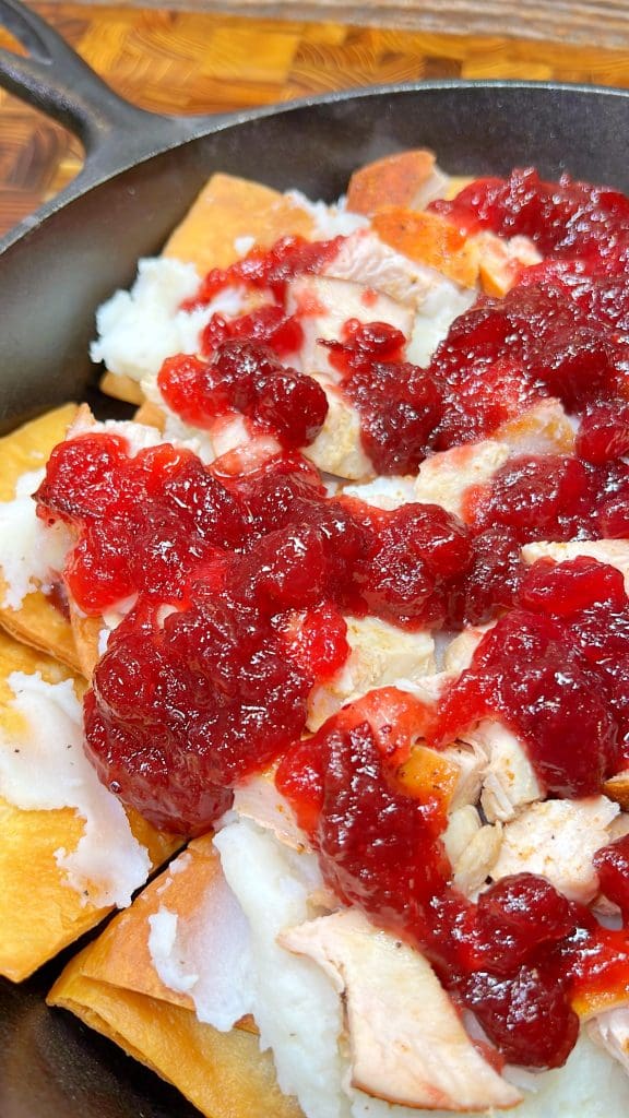 A close-up of a dish featuring crispy tortillas topped with shredded turkey, mashed potatoes, and cranberry sauce, served in a skillet. The vibrant red of the cranberry sauce contrasts with the lighter colors of the turkey and potatoes.