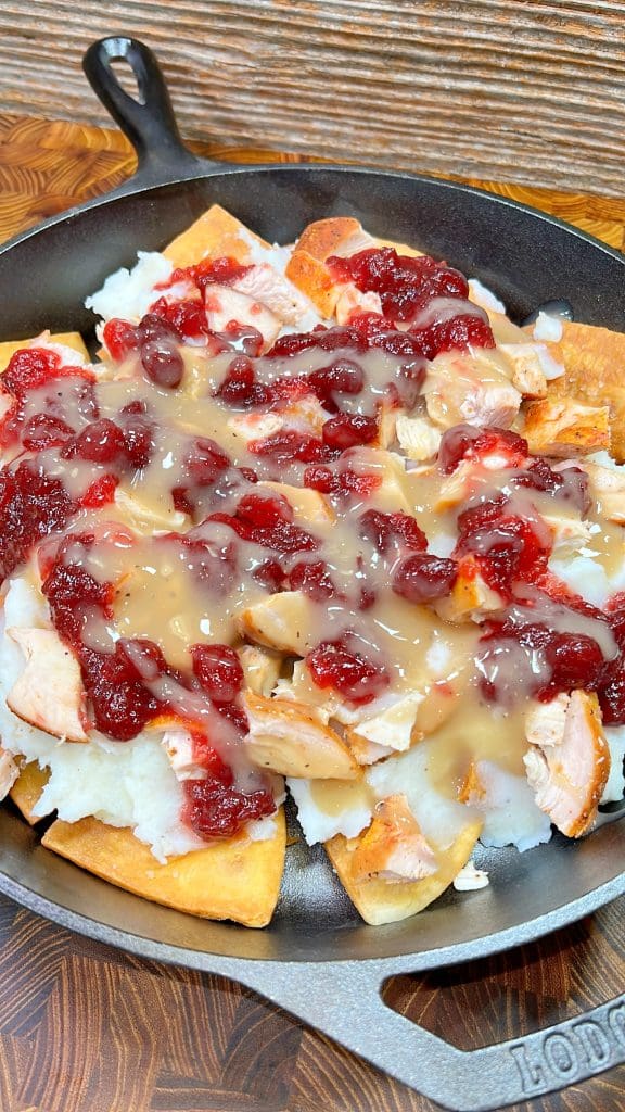 A skillet filled with a dish of turkey pieces, mashed potatoes, and flatbread, topped with cranberry sauce and gravy, sits on a wooden surface.