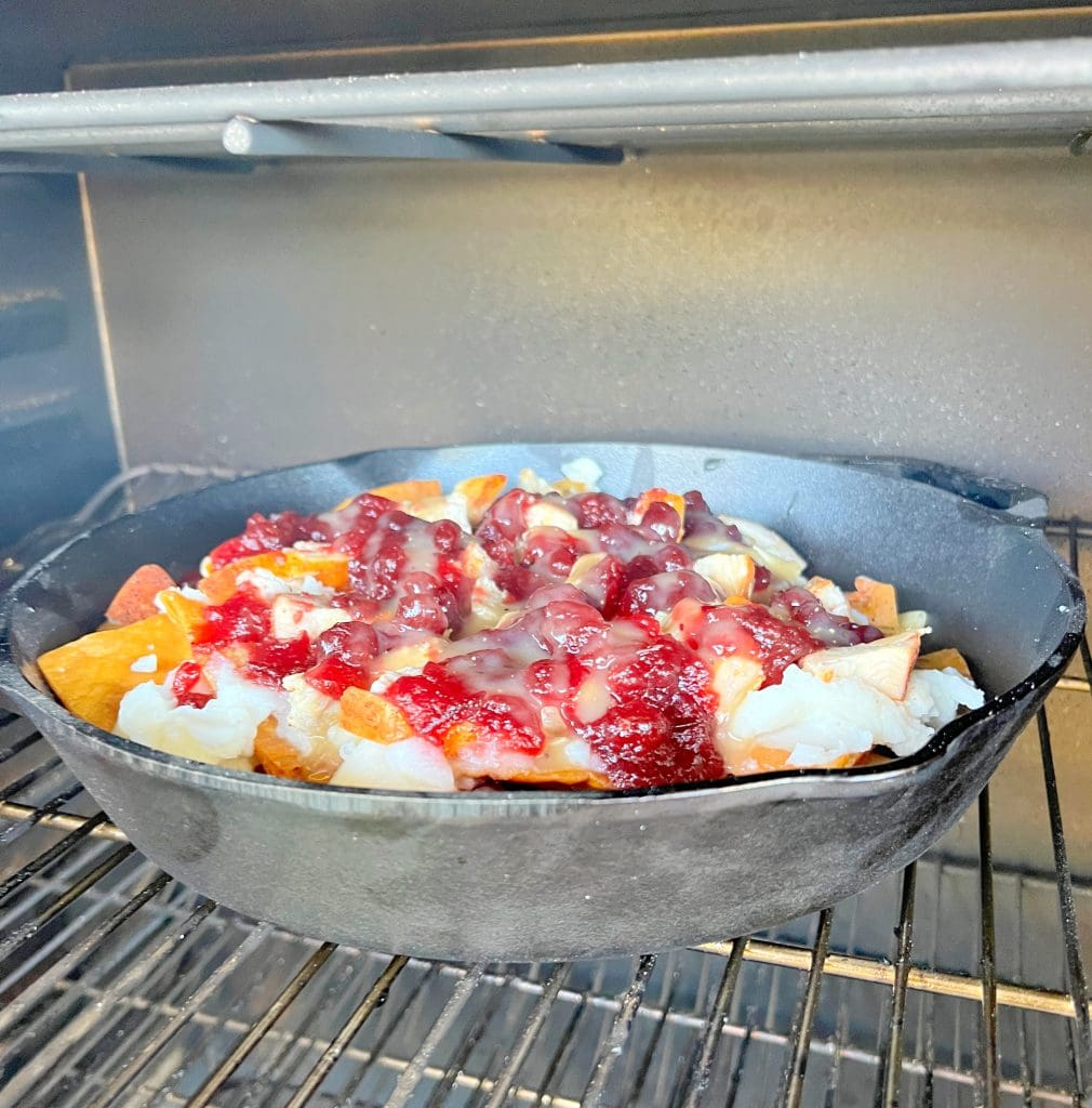 A cast iron skillet filled with nachos topped with melted cheese, diced onions, and a red sauce sits on a grill rack inside an oven.