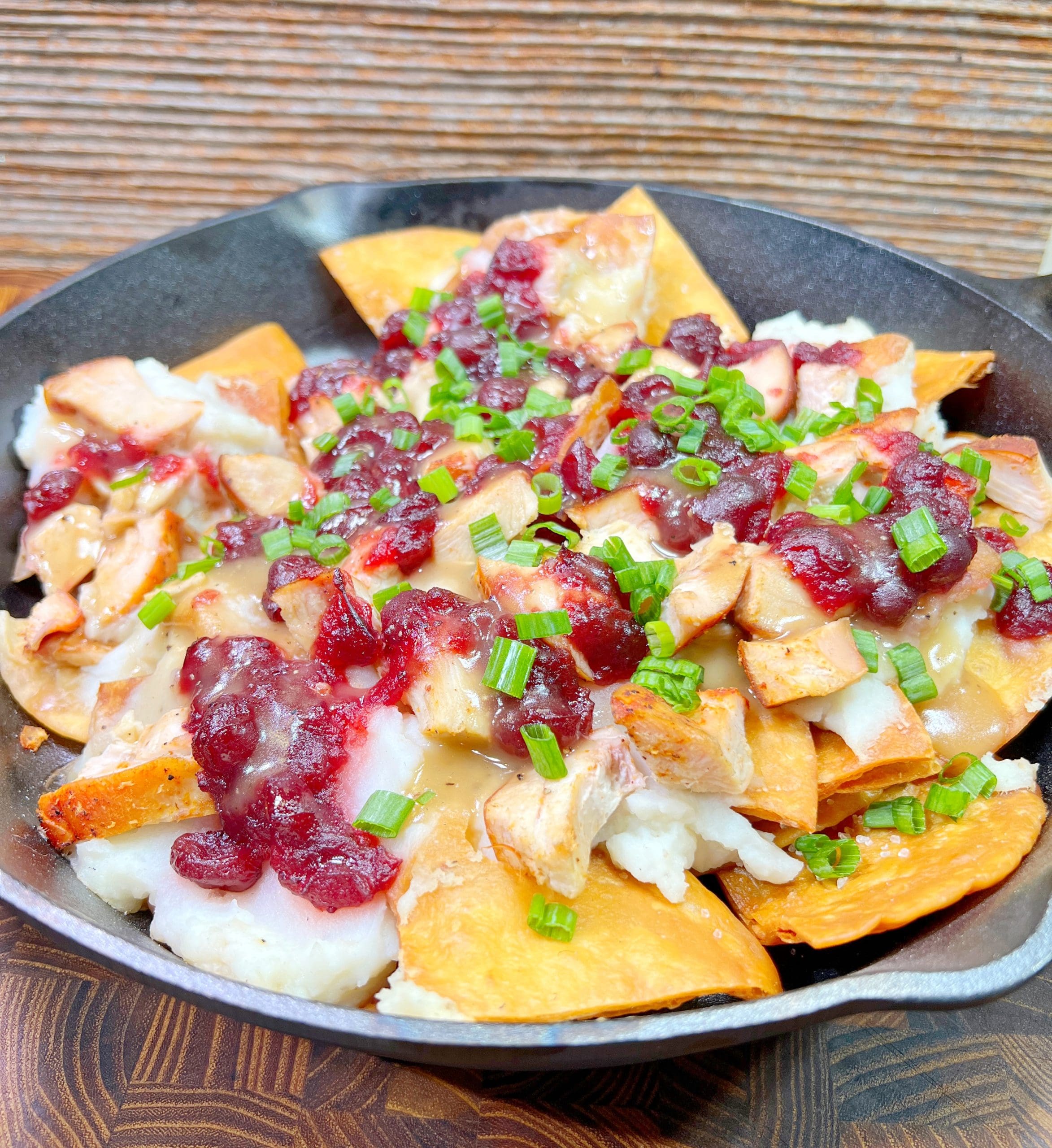 A skillet filled with a hearty dish of nachos topped with chunks of roasted turkey, gravy, cranberry sauce, and a sprinkle of chopped green onions. The background features a rustic wooden texture.