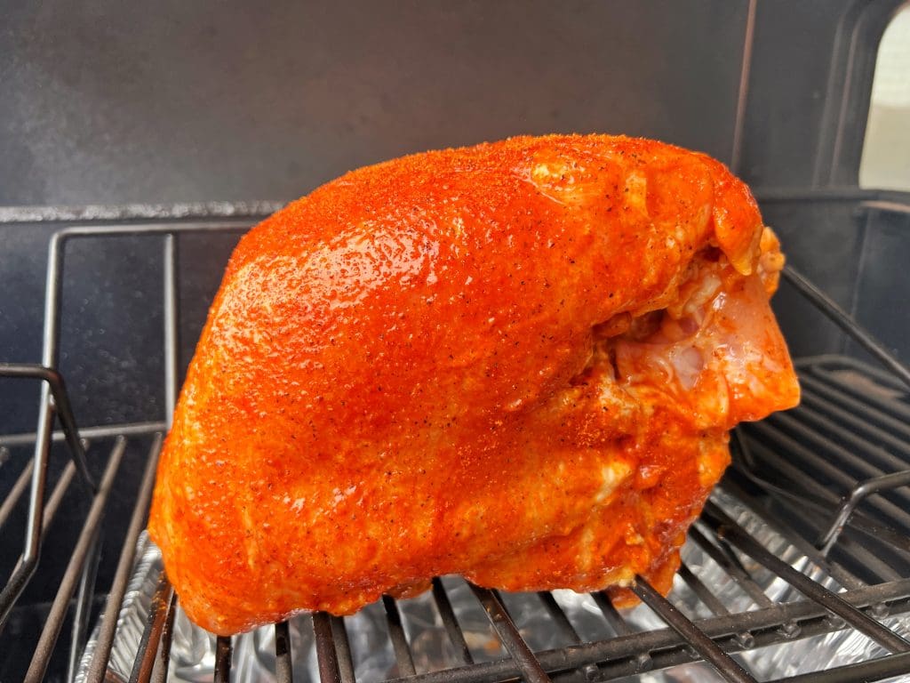 A seasoned turkey breast is placed on a grill, its surface coated with a rich red spice rub. The grills metal grates are visible beneath, and a foil tray catches any drippings.