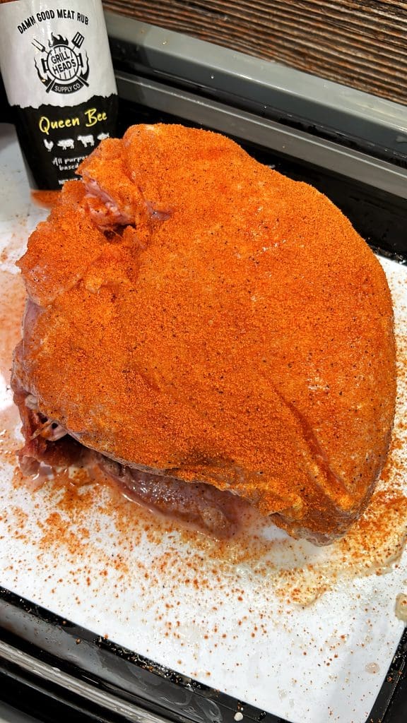 A large piece of raw meat on a tray is covered with a thick layer of orange seasoning. A bottle labeled Queen Bee from the brand Damn Good Meat Rub is visible in the background. The tray sits on a white surface.