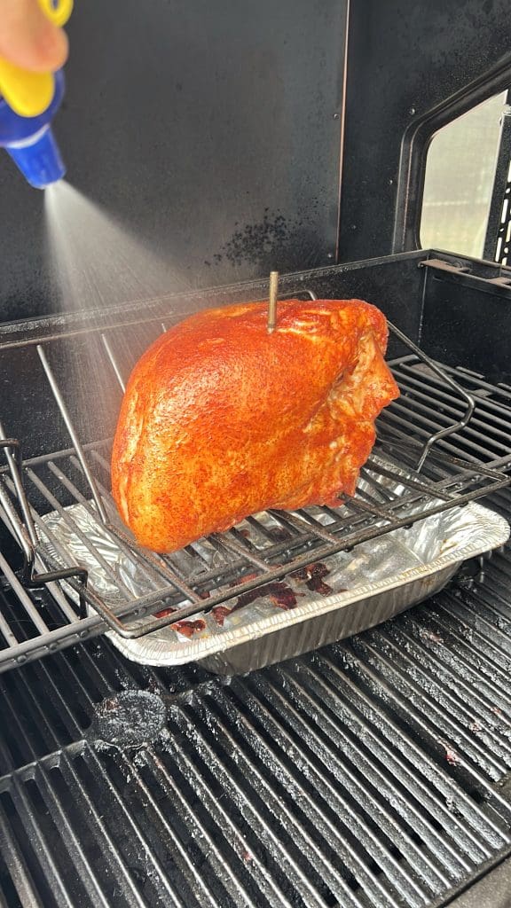 A large piece of seasoned meat is being smoked on a grill, with a metal probe inserted. A hand sprays liquid onto the meat for moisture. Below, a tray catches drippings. Smoke surrounds the grill area.