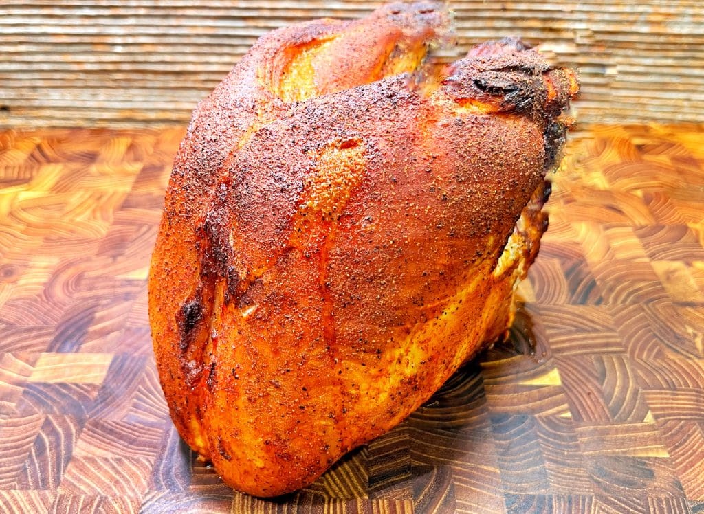 A seasoned and smoked turkey breast resting on a wooden cutting board with a textured background. The turkeys skin is golden brown and crispy, showcasing a well-cooked exterior.