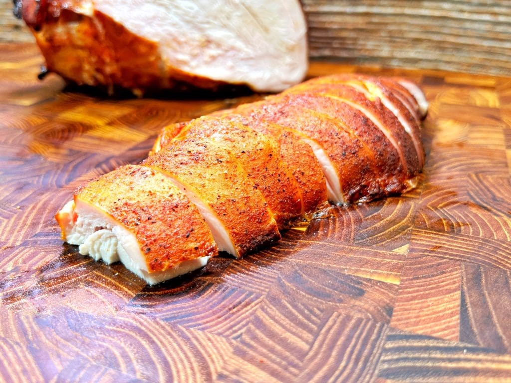 Sliced, roasted chicken breast on a wooden cutting board, with a whole roasted chicken in the background. The chicken skin is crispy and golden brown.