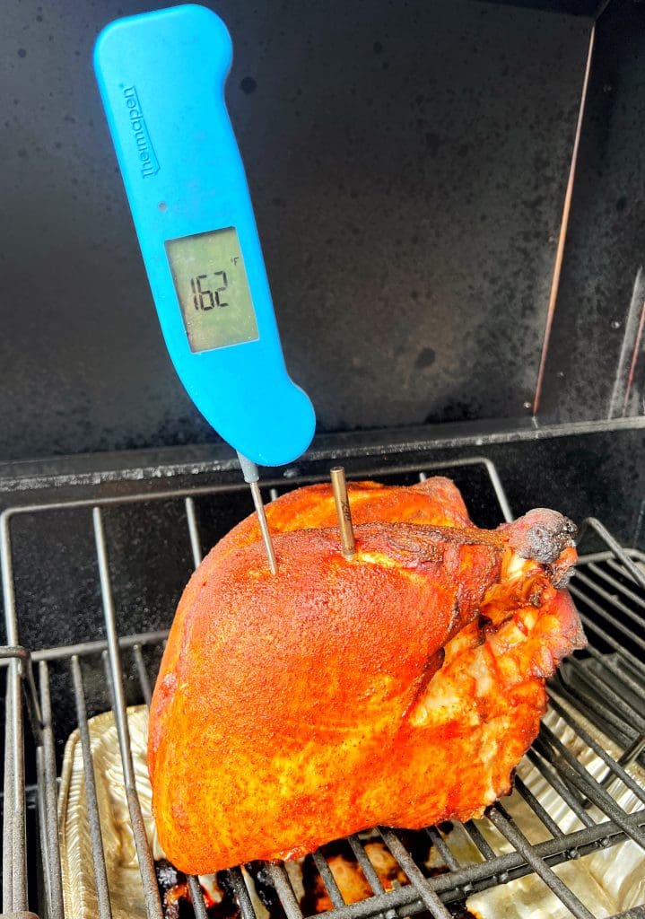 A seasoned piece of meat sits on a grill with a blue digital thermometer inserted into it, displaying a temperature of 162 degrees Fahrenheit. The grill has a smoky, blackened interior.