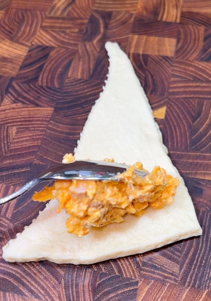 A triangular piece of dough on a wooden surface with a spoonful of orange filling placed in the center, ready for folding or rolling.