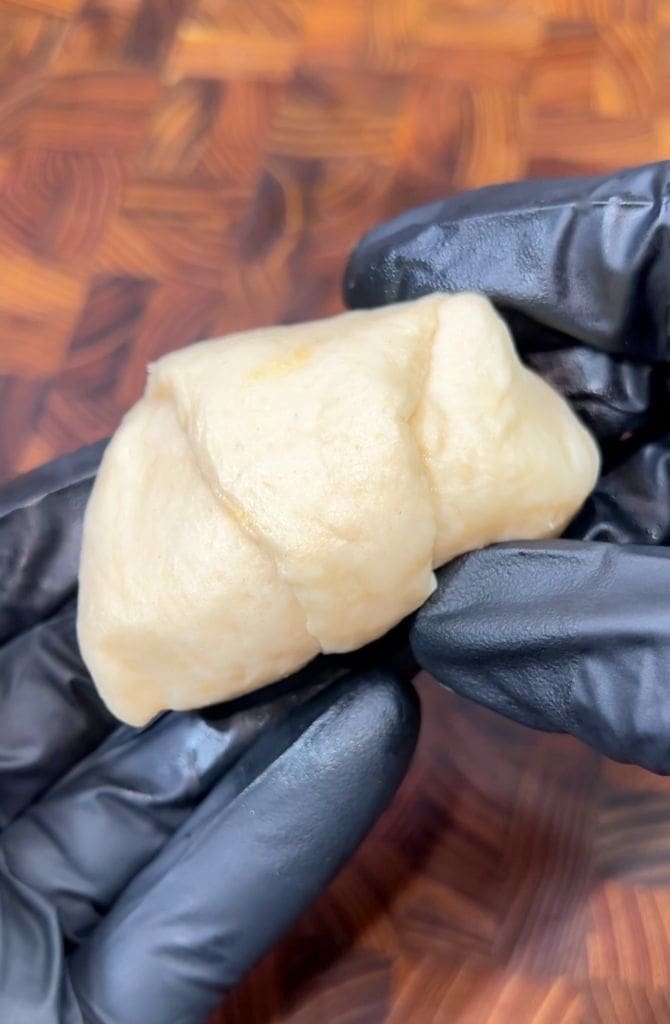 Gloved hands holding a small piece of dough against a wooden surface. The dough appears soft and smooth.