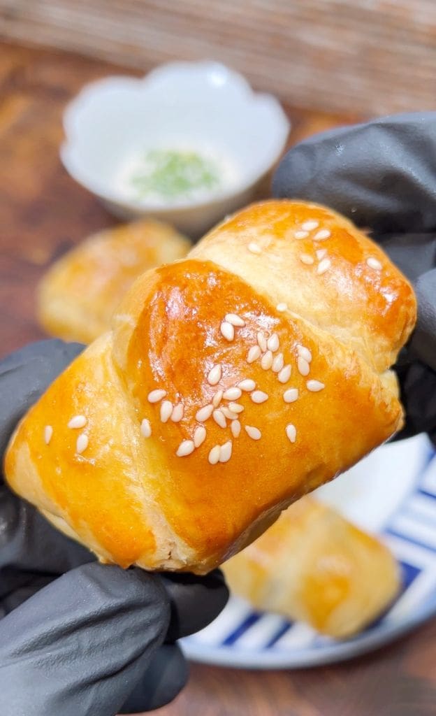 A hand with black gloves holds a golden-brown pastry topped with sesame seeds. Another pastry and a white bowl with green garnish are blurred in the background, all resting on a wooden surface.