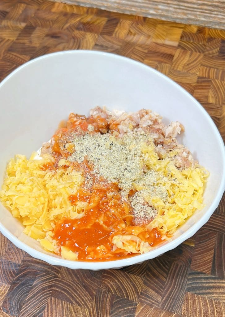 A white bowl on a wooden surface contains shredded cheddar cheese, cooked chicken, hot sauce, and seasoning.