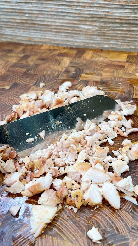 Chopped cooked chicken on a wooden cutting board with a large knife resting beside it. The board has a patterned surface, and the chicken pieces are small and scattered, showing a mixture of white and browned parts.