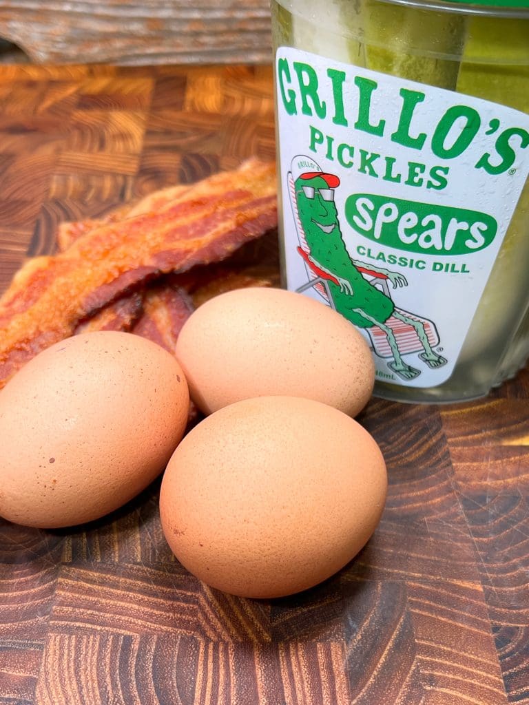 A jar of Grillos pickles, three brown eggs, and several strips of cooked bacon are arranged on a wooden surface. The pickles jar features a cartoon pickle wearing sunglasses and lounging on a chair.