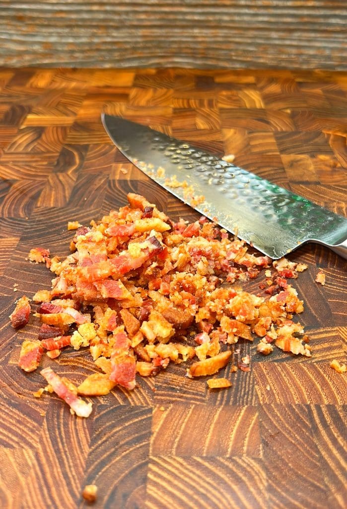 A sharp, textured knife sits on a wooden cutting board next to a pile of finely chopped bacon bits. The board has a rich, patterned grain, contrasting with the crispy, red-brown bacon pieces.