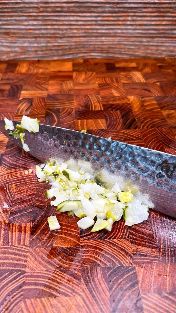 A close-up of chopped onions on a wooden cutting board with a large chefs knife visible. The board has a rich, patterned texture.