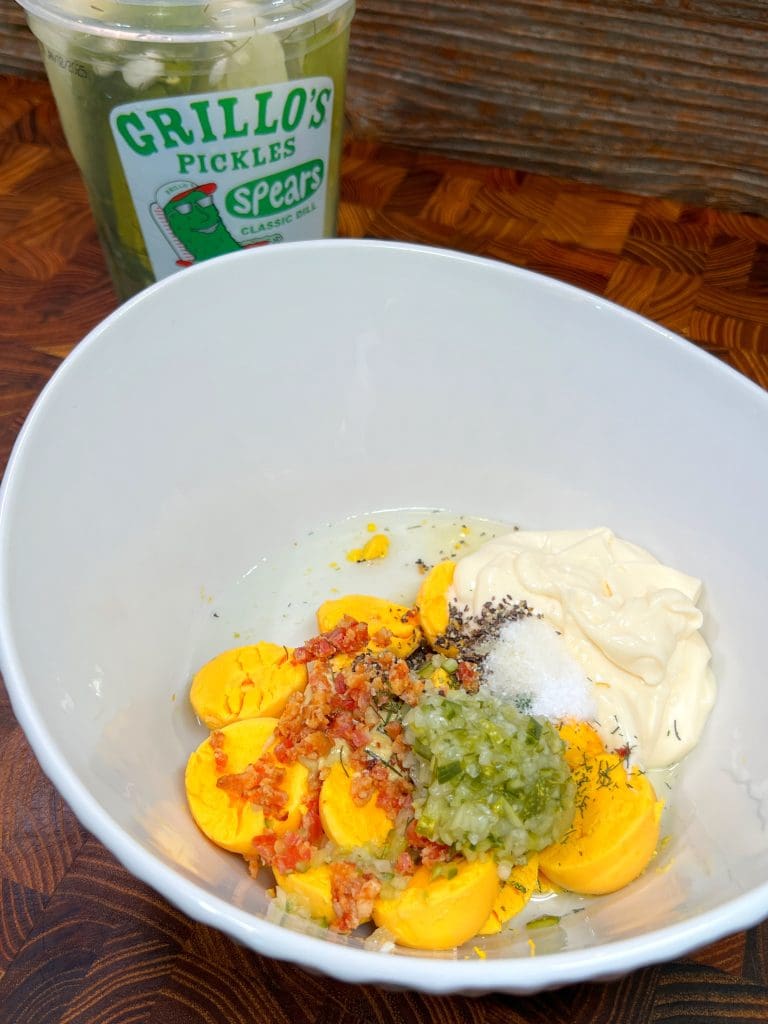 A white bowl contains egg yolks, mayonnaise, relish, bacon bits, and seasonings ready for mixing. In the background, there is a container of Grillos pickles on a wooden surface.