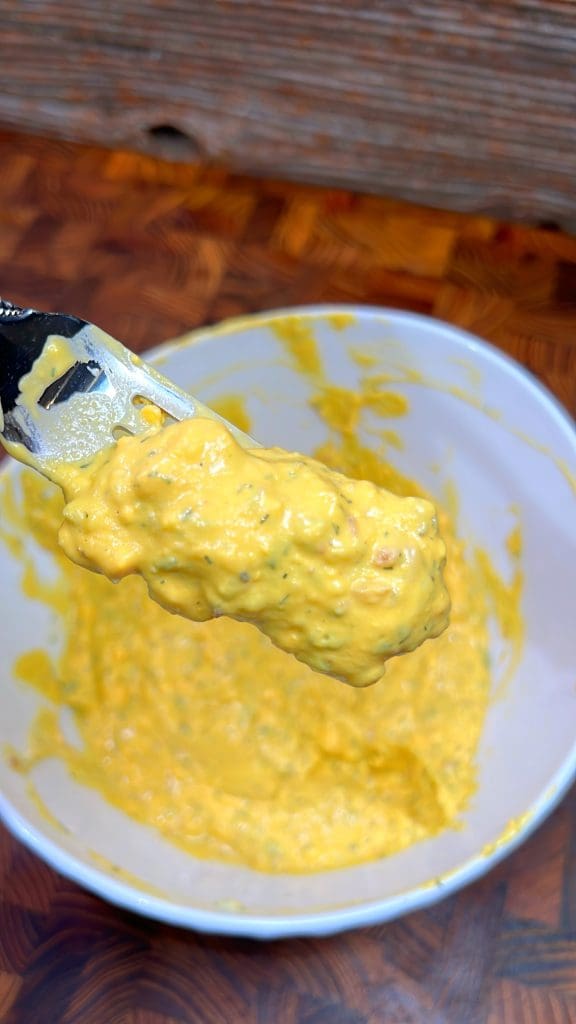 A spoon is lifting a thick, yellow batter with green flecks from a white bowl. The bowl is resting on a wooden surface with a textured pattern.