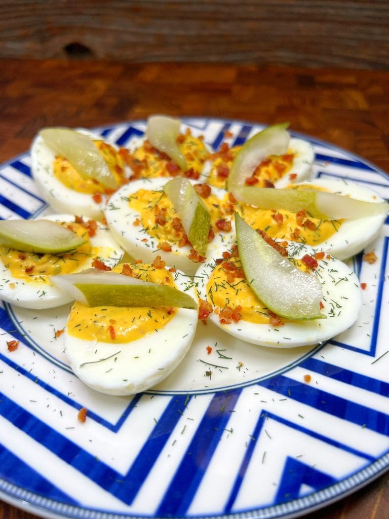 A plate of six deviled eggs, topped with crumbled bacon, dill, and pickle slices, arranged on a blue and white geometric-patterned plate. The eggs are set on a wooden surface.