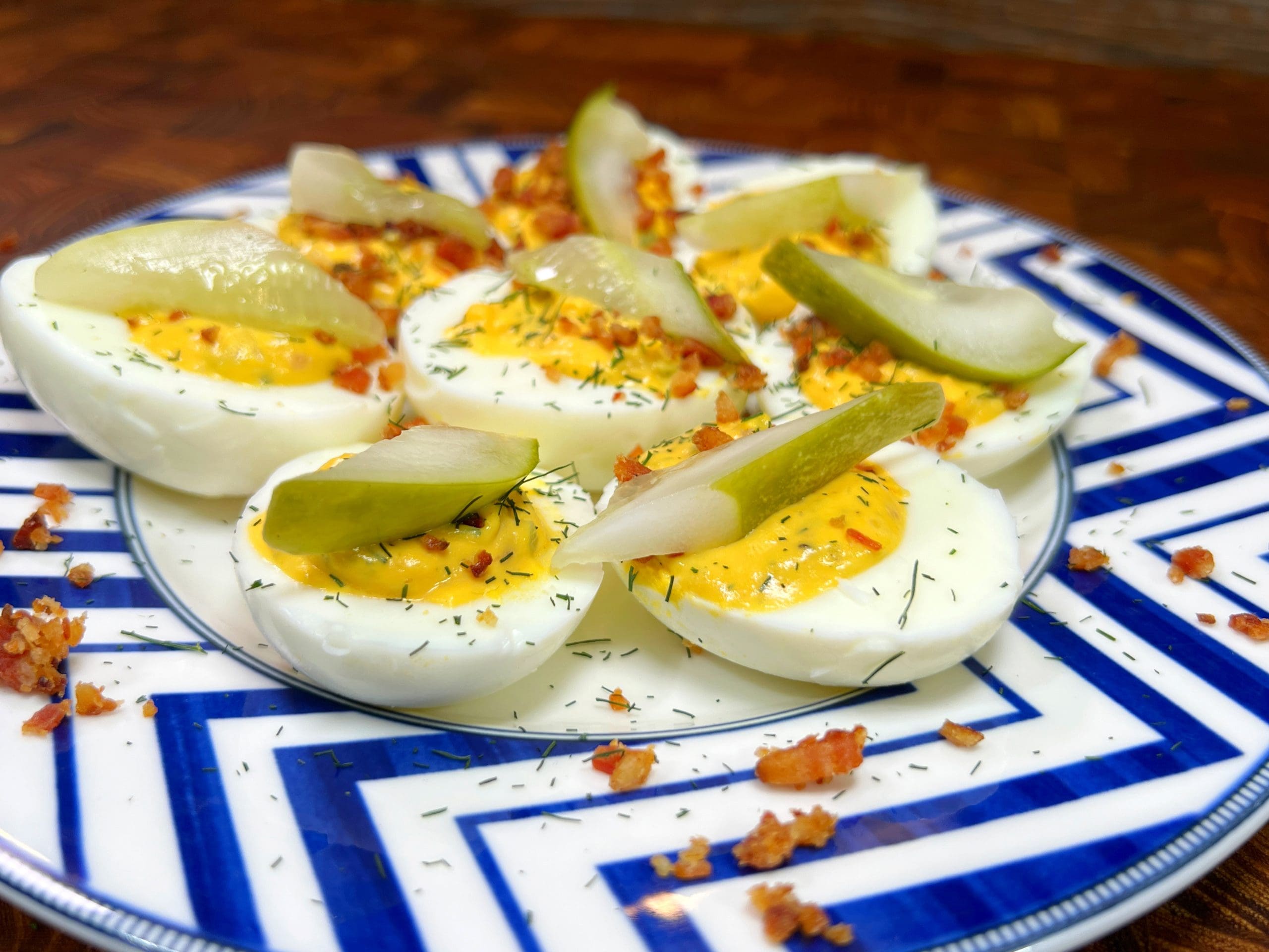 Deviled eggs topped with dill and pickle slices are served on a blue and white geometric-patterned plate. Crumbled bacon is scattered around the eggs, enhancing the presentation on a wooden table.