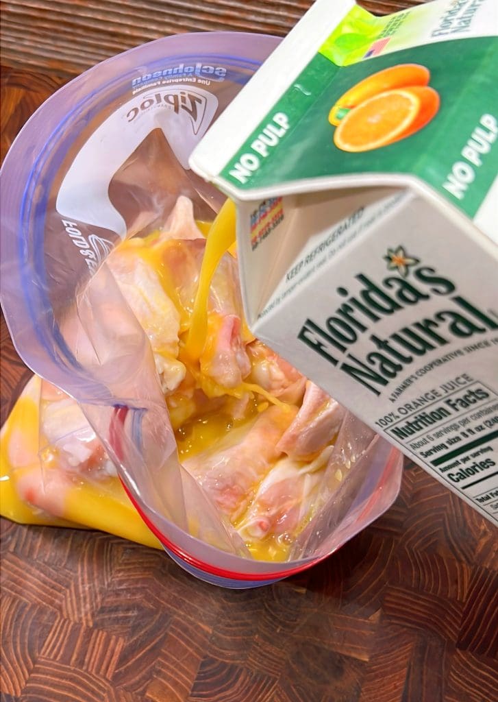 A carton of Floridas Natural orange juice is being poured into a resealable plastic bag containing raw chicken pieces. The bag is placed on a wooden surface.