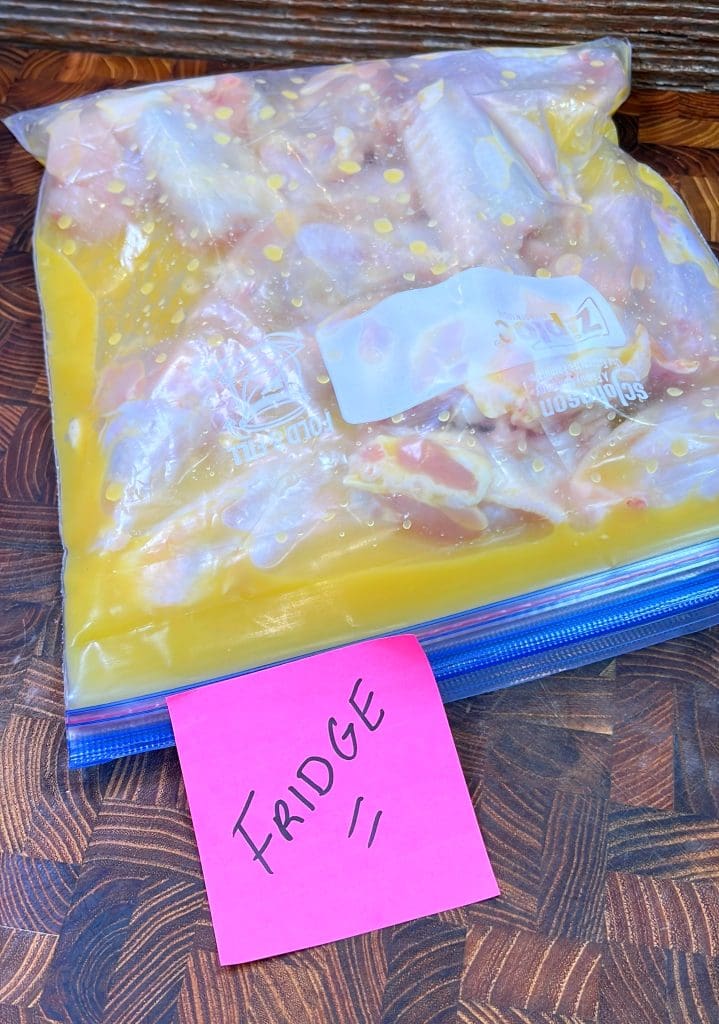 A ziplock bag filled with marinating chicken wings sits on a wooden surface. A pink sticky note labeled FRIDGE is placed in front of the bag.