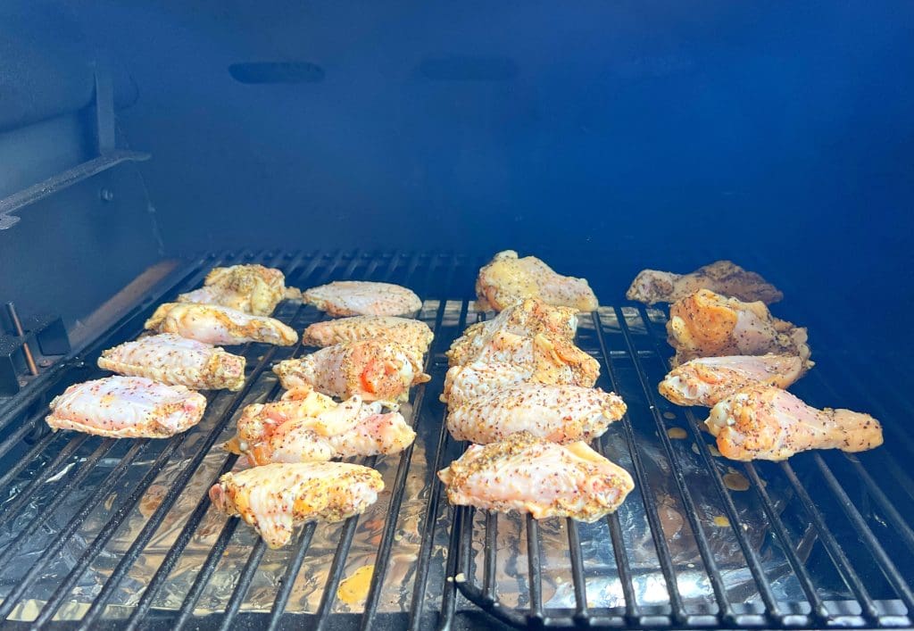 Chicken wings seasoned with pepper and spices are placed evenly on a grill, cooking over open flames. The grill marks are starting to show, and the surrounding smoke indicates they are being barbecued.