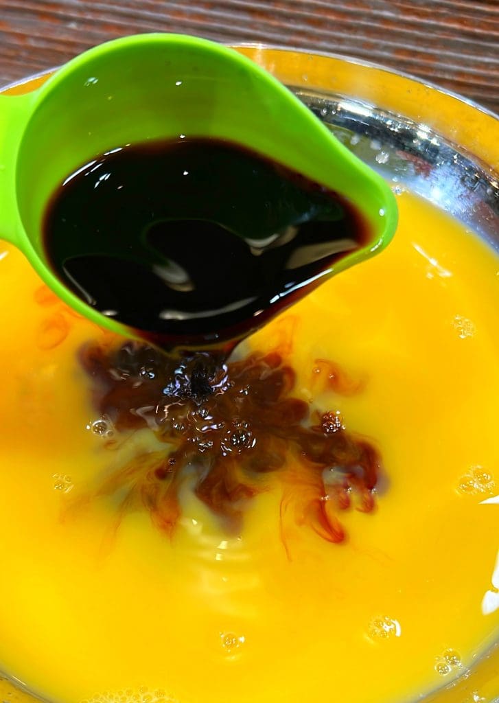 A green measuring cup pours dark soy sauce into a shiny metal bowl filled with beaten eggs, creating a swirling effect. The wooden surface beneath adds contrast to the vibrant colors of the ingredients.