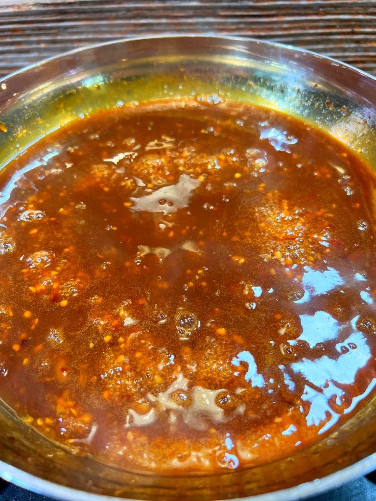 A close-up of a saucepan filled with a rich, bubbling brown sauce. The texture is smooth with visible flecks of spices, and the surface appears glossy. The pan is resting on a dark countertop.