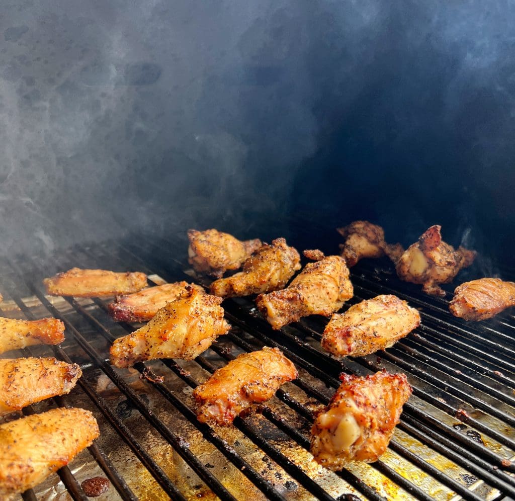 Chicken wings being grilled on a barbecue, with a smoky backdrop. The wings are seasoned, showcasing a crispy, golden-brown exterior. The smoke adds a rustic feel to the cooking process.