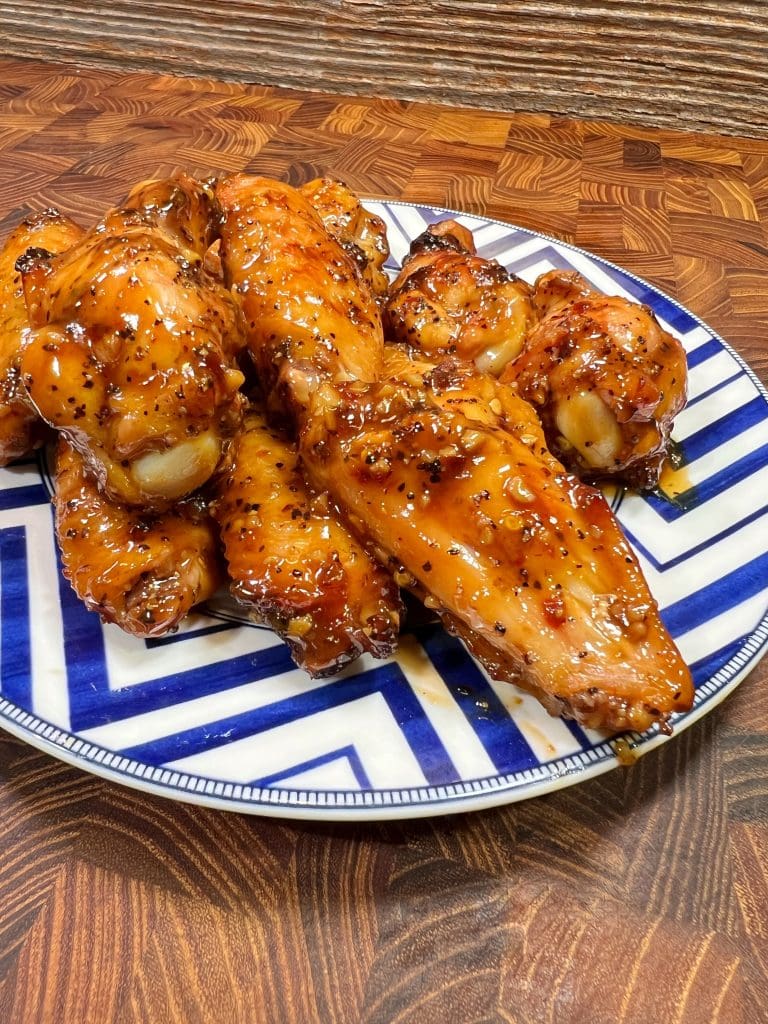 A chicken wings recipe with a plate of glazed chicken wings with a shiny, golden-brown coating on a blue and white chevron-patterned dish. The table has a wooden texture, enhancing the presentation of the dish.