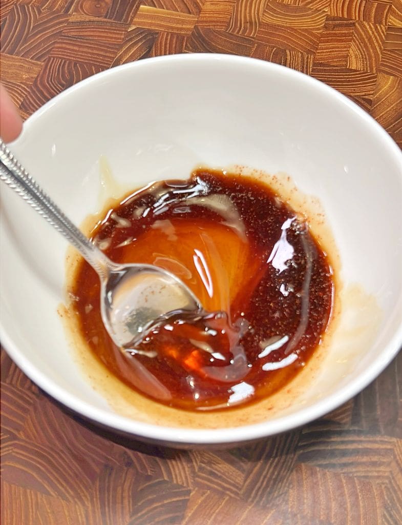 A white bowl on a wooden table contains a mixture of soy sauce and sugar. A silver spoon is partially submerged in the sticky, dark brown liquid, stirring the ingredients together.